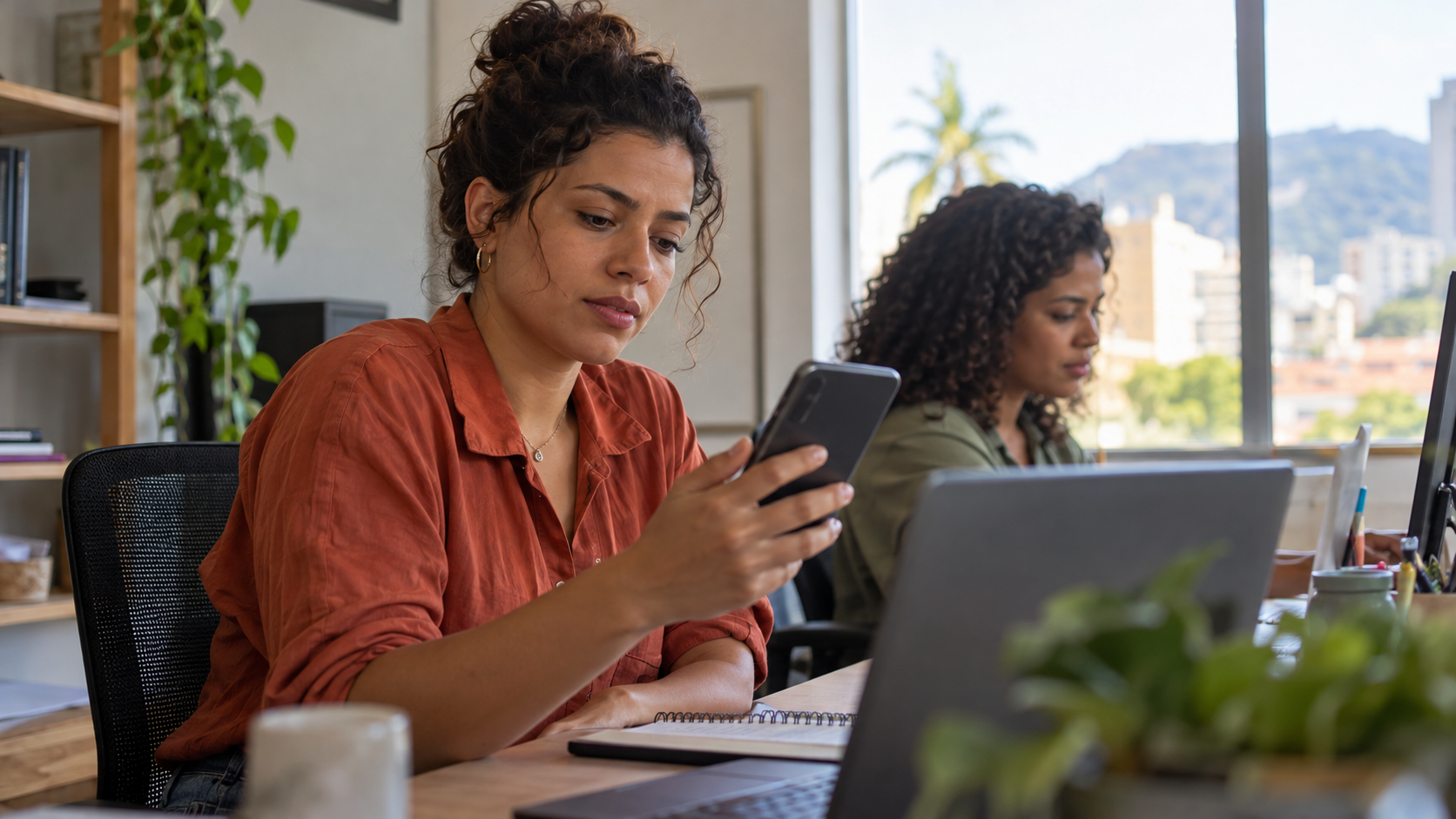 Atendente de uma pequena empresa lendo com atenção uma mensagem no celular, ao lado de uma colega, em um ambiente de trabalho comum