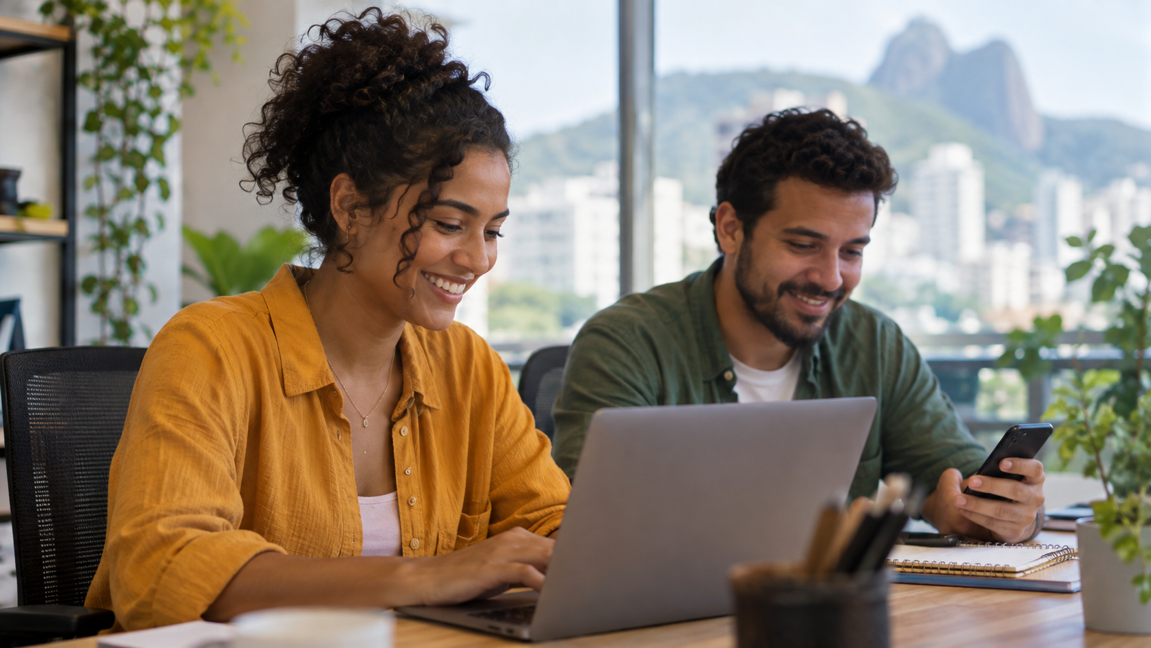 Duas pessoas de uma equipe de atendimento trabalhando juntas num escritório iluminado, uma delas respondendo uma mensagem no computador com um sorriso